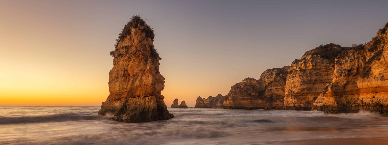 How To Visit Dona Ana Beach (Full Guide for 2026) 5 A stunning coastal landscape at sunset with rock formations and calm ocean waves at Praia Dona Ana