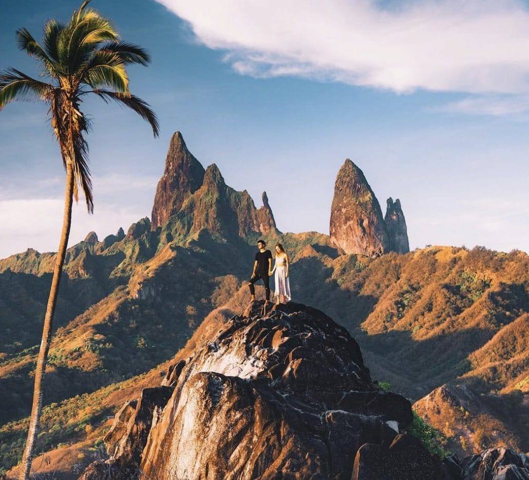 a man and woman standing on a rock with a palm tree and mountains in the background in Ua Pou!
