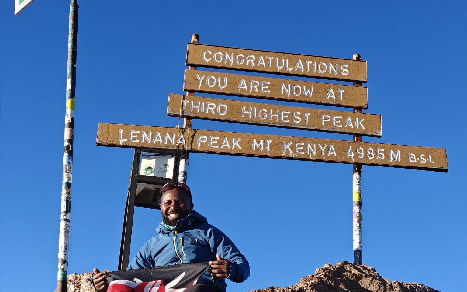 Mountain peak at Lenana Peak, Mount Kenya, with a hiker holding a Kenyan flag, celebrating reaching the third highest point at 4,985 meters above sea level under a clear blue sky.