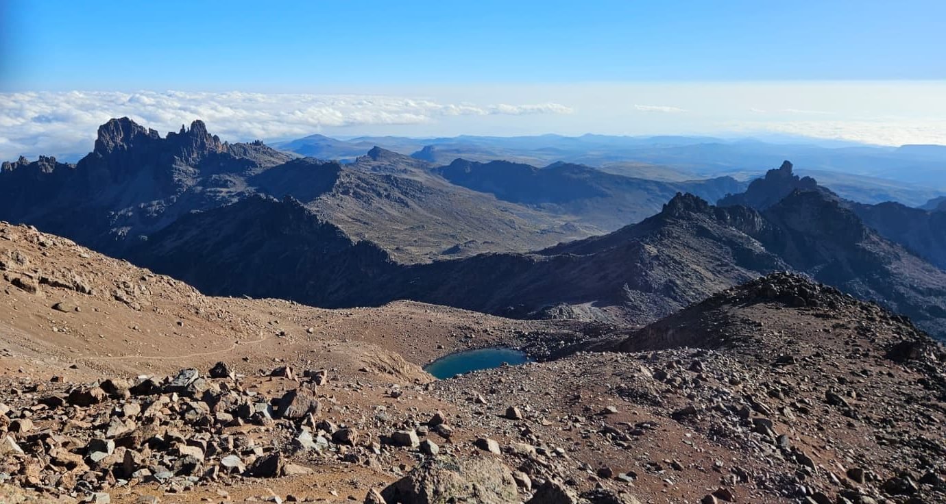 Vast mountain landscape at high altitude with rocky terrain, rugged peaks, and a small turquoise lake under a clear blue sky, capturing the breathtaking beauty of wilderness and adventure.