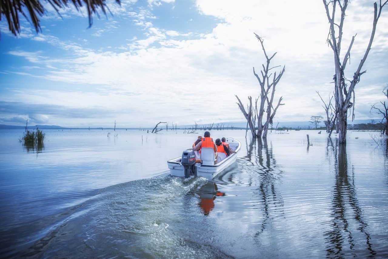 A boat with travelers navigating a flooded landscape at Lake Naivasha with leafless trees and cloudy skies, capturing the mysterious and atmospheric scenery of Lost Between Oceans.