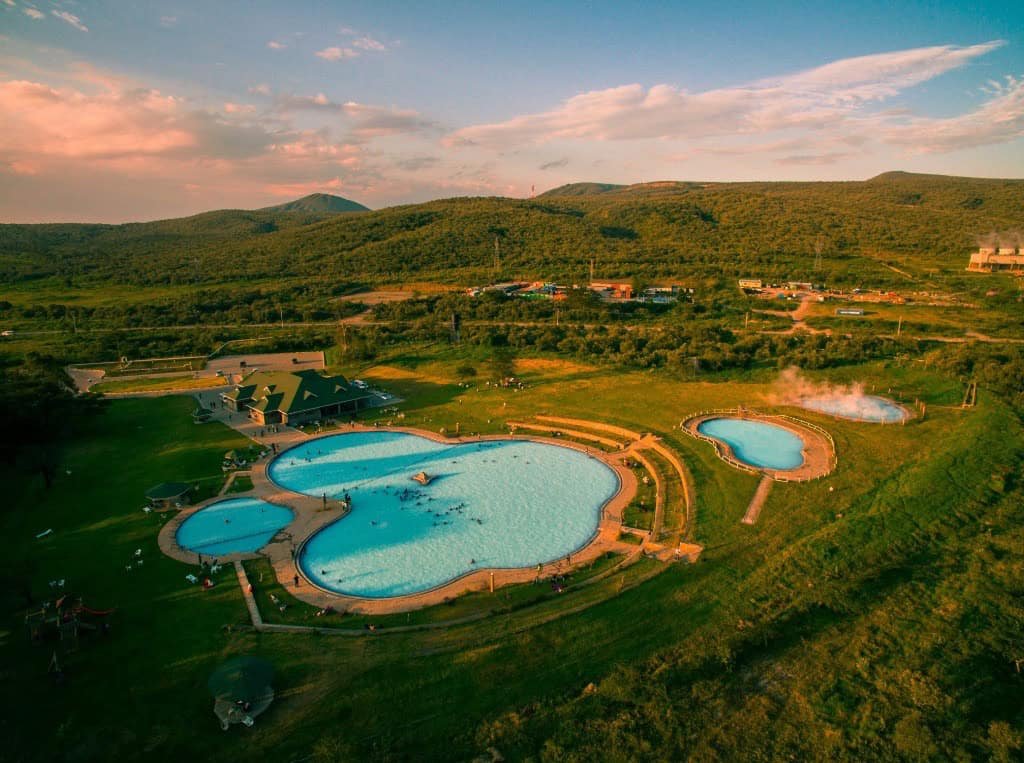 1. Aerial view of a scenic outdoor swimming pool complex surrounded by lush green hills and mountains at sunset.